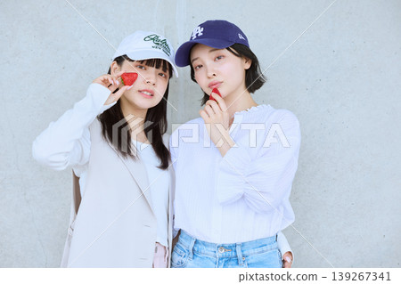 Two young women in caps posing with strawberries - a stylish street snap of friends. Two young women in caps posing with strawberries - a stylish street snap of friends. 139267341