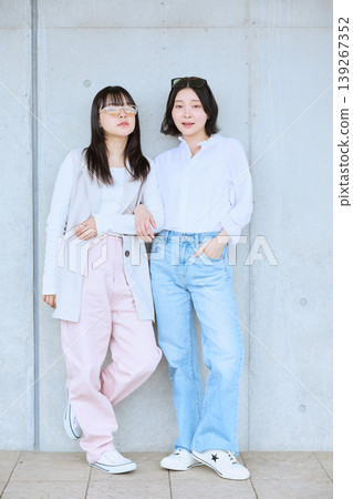 兩名年輕女子並肩站在一棟混凝土建築前;身著休閒春裝;與朋友外出。 兩名年輕女子並肩站在一棟混凝土建築前;身著休閒春裝;與朋友外出。 139267352