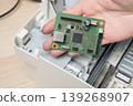 Technician holding a green circuit board inside an open printer, inspecting electronic components for repair and maintenance. PCB chips, connectors and hands-on troubleshooting in a workshop setting. 139268907