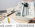 A technician services a disassembled office printer, exposing rollers, circuit boards and cables during maintenance and troubleshooting to repair paper feed and electronic components. 139268908
