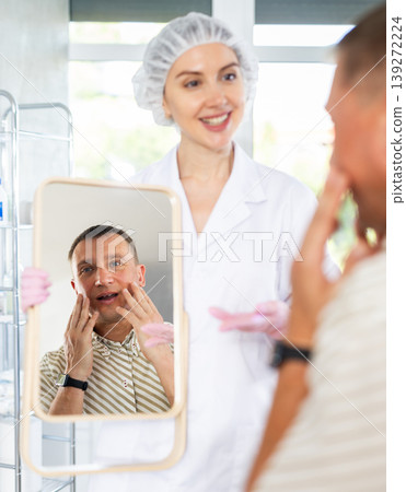 Nurse holds a mirror, satisfied man examines his face in mirror after plastic surgery Nurse holds a mirror, satisfied man examines his face in mirror after plastic surgery 139272224