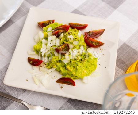 Pile of guacamole on plate, sprinkled with chopped hot onions, decorated with slices of tomatoes 139272249