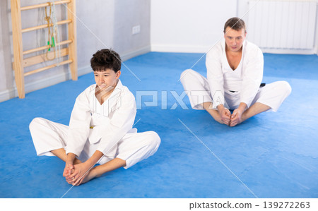 Teenage boy and middle-aged man trainer wearing kimono doing leg stretching in sports hall Teenage boy and middle-aged man trainer wearing kimono doing leg stretching in sports hall 139272263
