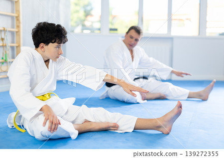 Teenage boy and middle-aged man trainer wearing kimono doing leg stretching in sports hall 139272355