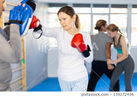 Woman in boxing gloves practices punches with trainer in mitts in sport club during family training 139272436