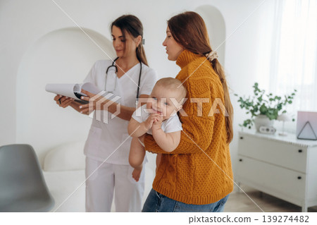 Information being written into notepad. Mother with her little baby daughter is in clinic with doctor at appointment Information being written into notepad. Mother with her little baby daughter is in clinic with doctor at appointment 139274482