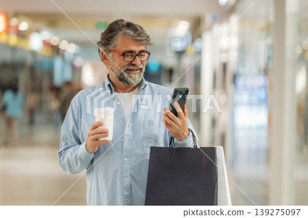 Cup of drink and smartphone are in hands. Senior man is in the mall, shopping time conception Cup of drink and smartphone are in hands. Senior man is in the mall, shopping time conception 139275097