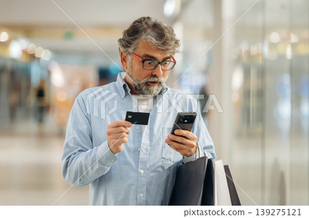 Very shocked, holding credit or debit card and smartphone. Senior man is in the mall, shopping time conception 139275121