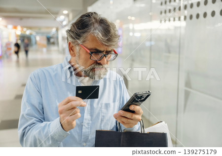 Funny facial expression, holding credit card and smartphone. Senior man is in the mall, shopping time conception Funny facial expression, holding credit card and smartphone. Senior man is in the mall, shopping time conception 139275179