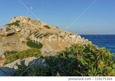 A serene coastal view with flags atop a rocky hill by the azure sea during a sunny afternoon. A serene coastal view with flags atop a rocky hill by the azure sea during a sunny afternoon. 139275368