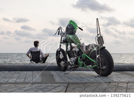 Perched upon a folding chair, a solitary man looks toward the sea, his back to a custom green motorcycle that stands as his mechanical companion. The bike striking mohawk helmet offers a rebellious Perched upon a folding chair, a solitary man looks toward the sea, his back to a custom green motorcycle that stands as his mechanical companion. The bike striking mohawk helmet offers a rebellious 139275621