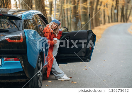 Door is opened, sitting and holding drink. Beautiful blonde woman is with her blue electric car on the road of forest Door is opened, sitting and holding drink. Beautiful blonde woman is with her blue electric car on the road of forest 139275652