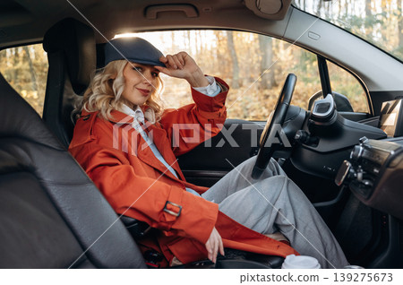 In red coat and blue beret. Happy blonde woman is sitting her electric car In red coat and blue beret. Happy blonde woman is sitting her electric car 139275673