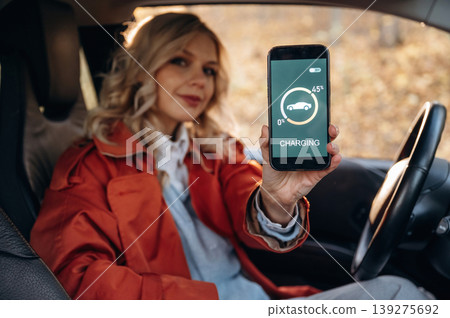 App in smartphone shows that automobile is charging. Happy blonde woman is sitting her electric car 139275692