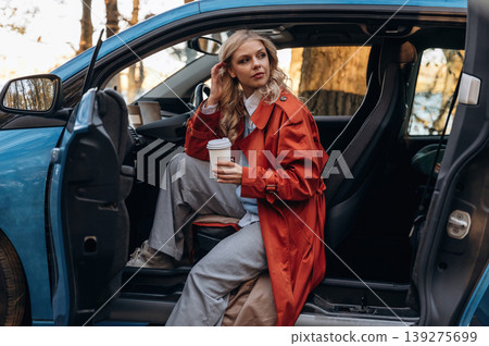 Side view, with cup of drink. Beautiful blonde woman is with her blue electric car on the road of forest Side view, with cup of drink. Beautiful blonde woman is with her blue electric car on the road of forest 139275699