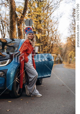 Side view, with cup of drink. Beautiful blonde woman is with her blue electric car on the road of forest Side view, with cup of drink. Beautiful blonde woman is with her blue electric car on the road of forest 139275700