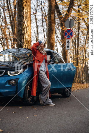 Leaning on the automobile, holding cup of drink. Beautiful blonde woman is with her blue electric car on the road of forest Leaning on the automobile, holding cup of drink. Beautiful blonde woman is with her blue electric car on the road of forest 139275701