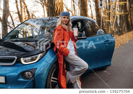 Leaning on the automobile, holding cup of drink. Beautiful blonde woman is with her blue electric car on the road of forest Leaning on the automobile, holding cup of drink. Beautiful blonde woman is with her blue electric car on the road of forest 139275704