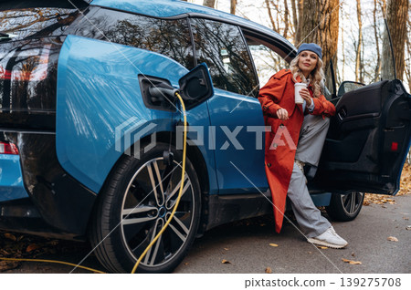 Automobile is charging. Beautiful blonde woman is with her blue electric car on the road of forest Automobile is charging. Beautiful blonde woman is with her blue electric car on the road of forest 139275708