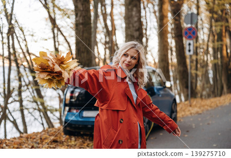 Bunch of fallen leaves, fun. Beautiful blonde woman is with her blue electric car on the road of forest 139275710