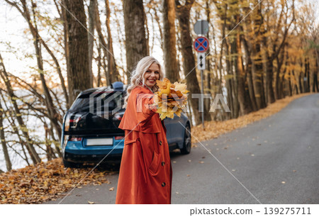 Bunch of fallen leaves, fun. Beautiful blonde woman is with her blue electric car on the road of forest 139275711