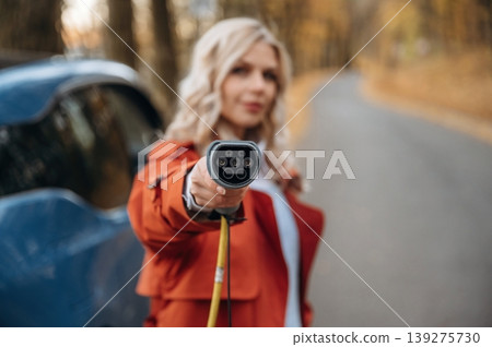 Holding the charging cable. Beautiful blonde woman is with her blue electric car on the road of forest 139275730