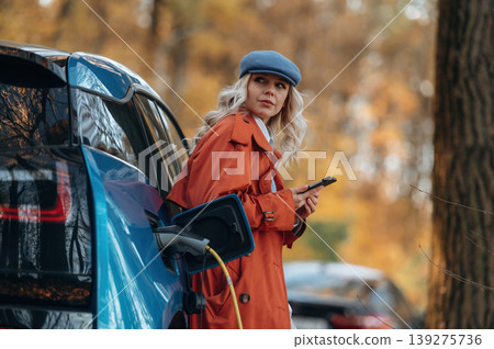 Black smartphone in hands, standing and waiting for charging to complete. Beautiful blonde woman is with her blue electric car on the road of forest Black smartphone in hands, standing and waiting for charging to complete. Beautiful blonde woman is with her blue electric car on the road of forest 139275736