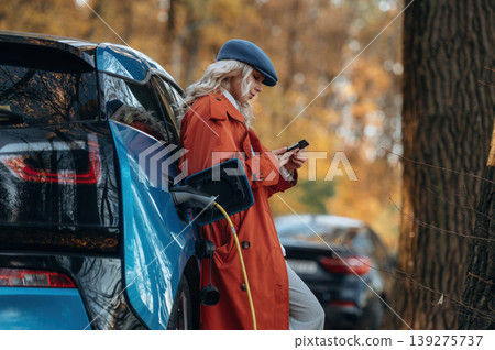 Black smartphone in hands, standing and waiting for charging to complete. Beautiful blonde woman is with her blue electric car on the road of forest Black smartphone in hands, standing and waiting for charging to complete. Beautiful blonde woman is with her blue electric car on the road of forest 139275737