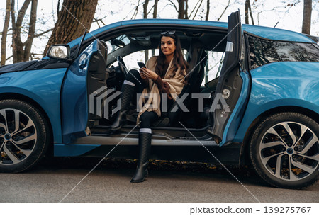 On the road, door opened, parked. Beautiful woman is sitting in her electric car On the road, door opened, parked. Beautiful woman is sitting in her electric car 139275767