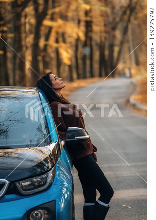 In brown sweater and black pants. Woman is outdoors near her electric blue car at daytime, rural scene In brown sweater and black pants. Woman is outdoors near her electric blue car at daytime, rural scene 139275792