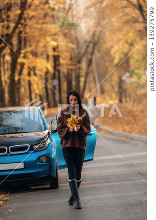 Sideroad, parked, having a walk. Woman is outdoors near her electric blue car at daytime, rural scene Sideroad, parked, having a walk. Woman is outdoors near her electric blue car at daytime, rural scene 139275799