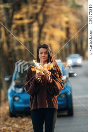 Against the vehicle that is parked behind. Woman is outdoors near her electric blue car at daytime, rural scene Against the vehicle that is parked behind. Woman is outdoors near her electric blue car at daytime, rural scene 139275816