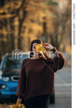 Leaves are in hands. Woman is outdoors near her electric blue car at daytime, rural scene Leaves are in hands. Woman is outdoors near her electric blue car at daytime, rural scene 139275820