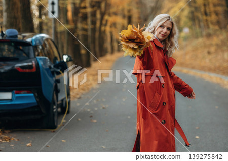 Bunch of fallen leaves, fun. Beautiful blonde woman is with her blue electric car on the road of forest Bunch of fallen leaves, fun. Beautiful blonde woman is with her blue electric car on the road of forest 139275842