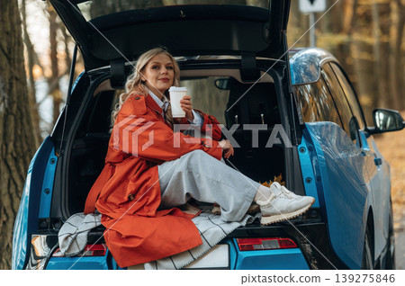 Sitting in the trunk. Beautiful blonde woman is with her blue electric car on the road of forest Sitting in the trunk. Beautiful blonde woman is with her blue electric car on the road of forest 139275846