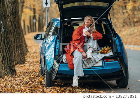 Cup of drink, sitting in the trunk. Beautiful blonde woman is with her blue electric car on the road of forest 139275851