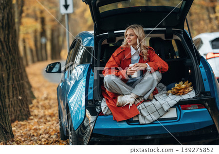Cup of drink, sitting in the trunk. Beautiful blonde woman is with her blue electric car on the road of forest 139275852