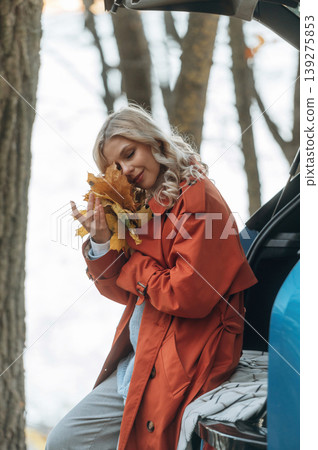 Having fun with fallen leaves. Beautiful blonde woman is with her blue electric car on the road of forest 139275853