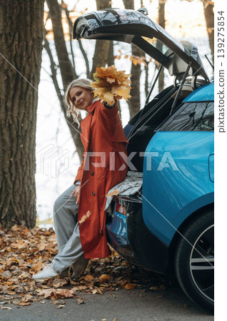 Having fun with fallen leaves. Beautiful blonde woman is with her blue electric car on the road of forest Having fun with fallen leaves. Beautiful blonde woman is with her blue electric car on the road of forest 139275854