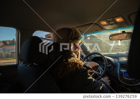 Man wearing winter hat driving a car. Caucasian driver commuting to work during cold season. Passenger perspective of taxicab service or delivery courier driving vehicle in morning light. Man wearing winter hat driving a car. Caucasian driver commuting to work during cold season. Passenger perspective of taxicab service or delivery courier driving vehicle in morning light. 139276259