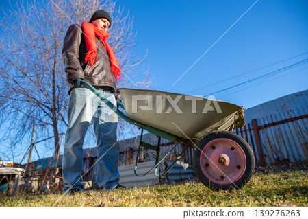 Man holding empty wheelbarrow in garden during autumn. Gardener preparing for yard work. Seasonal outdoor maintenance, land improvement and landscaping concept on sunny day. Man holding empty wheelbarrow in garden during autumn. Gardener preparing for yard work. Seasonal outdoor maintenance, land improvement and landscaping concept on sunny day. 139276263
