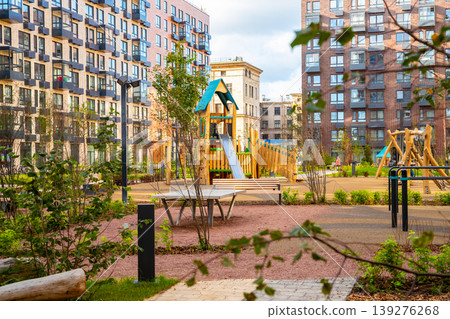 Modern urban playground courtyard between residential brick apartment buildings. Neighborhood outdoor recreational space with park bench, ping pong table and wooden climbing frame structure. Modern urban playground courtyard between residential brick apartment buildings. Neighborhood outdoor recreational space with park bench, ping pong table and wooden climbing frame structure. 139276268