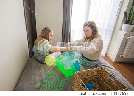 A girl and a woman work together to arrange a green hamster cage on a bed. A girl and a woman work together to arrange a green hamster cage on a bed. 139276533