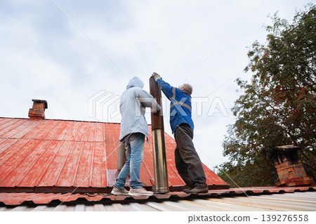 Two men installing metal chimney pipe on house roof. Construction workers performing roof maintenance and chimney repair. Professional renovation service of heating system in residence. Two men installing metal chimney pipe on house roof. Construction workers performing roof maintenance and chimney repair. Professional renovation service of heating system in residence. 139276558
