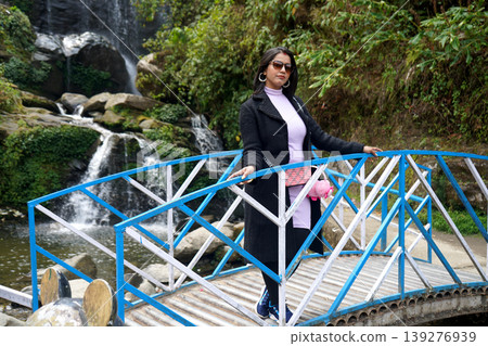 Stylish young woman in a black coat posing on a blue bridge with a scenic waterfall background at Rock Garden Darjeeling 139276939