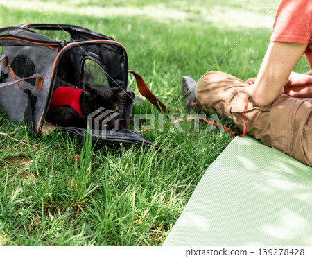 man sitting on grass with black cat in pet carrier. Person relaxing at park on picnic mat with domestic animal. Travel adventure with feline and pet supply for outdoor lifestyle. man sitting on grass with black cat in pet carrier. Person relaxing at park on picnic mat with domestic animal. Travel adventure with feline and pet supply for outdoor lifestyle. 139278428