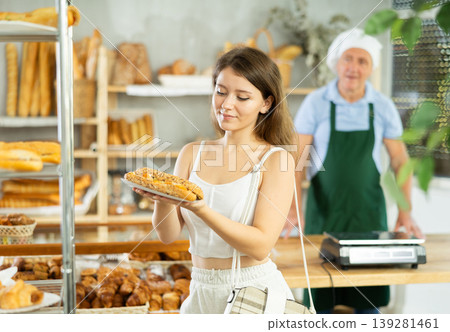 Positive beautiful girl buyer choosing and holding bocadillo in hands, elderly man seller standing in background in bakery shop Positive beautiful girl buyer choosing and holding bocadillo in hands, elderly man seller standing in background in bakery shop 139281461