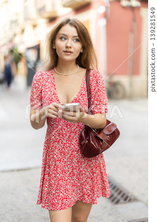 Girl in summer dress walks along street of the old town with mobile phone in her hands Girl in summer dress walks along street of the old town with mobile phone in her hands 139281549