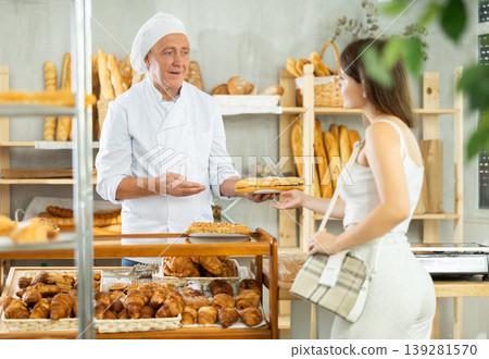 Positive mature man baker in white uniform offering to young girl fresh bocadillo in bakery Positive mature man baker in white uniform offering to young girl fresh bocadillo in bakery 139281570