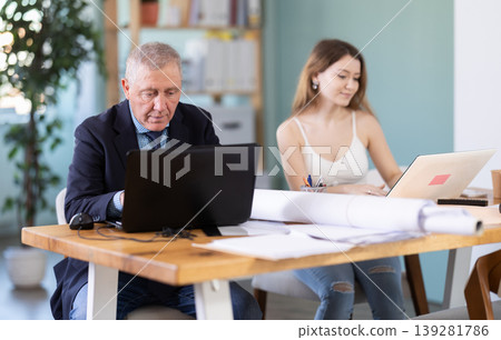Mature man working in office, sitting at desk next to woman Mature man working in office, sitting at desk next to woman 139281786